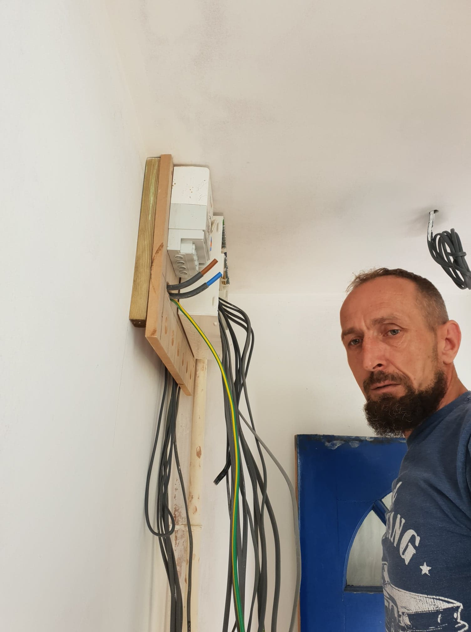 Rozdzielnia elektryczna zamontowana na drewnianej płycie przy białej ścianie. Widoczne kable i fragment twarzy elektryka. Błękitne drzwi w tle.