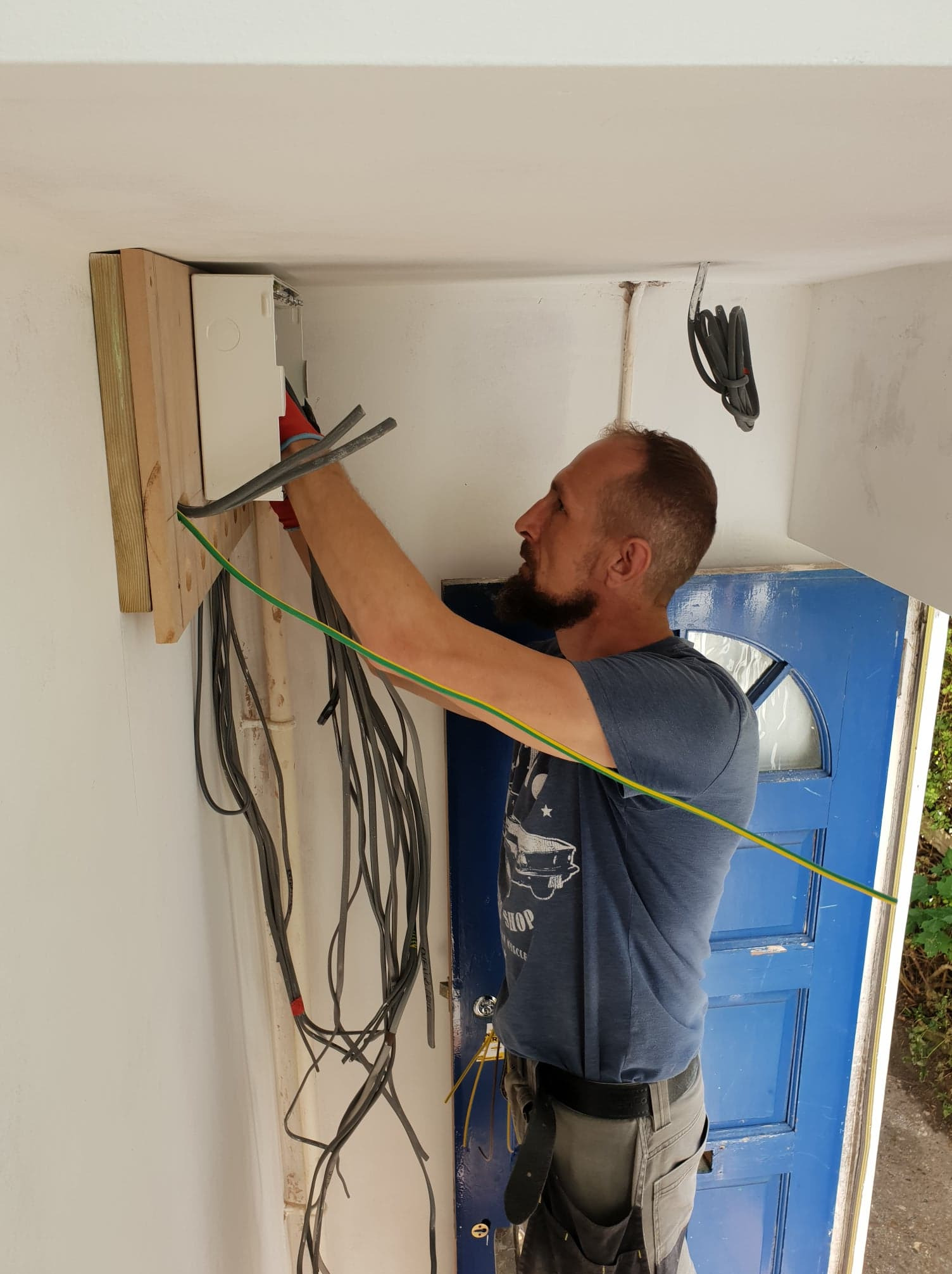 Elektryk w trakcie montażu skrzynki elektrycznej z licznymi kablami w pomieszczeniu z niebieskimi drzwiami. Widoczny etap prac instalacyjnych.