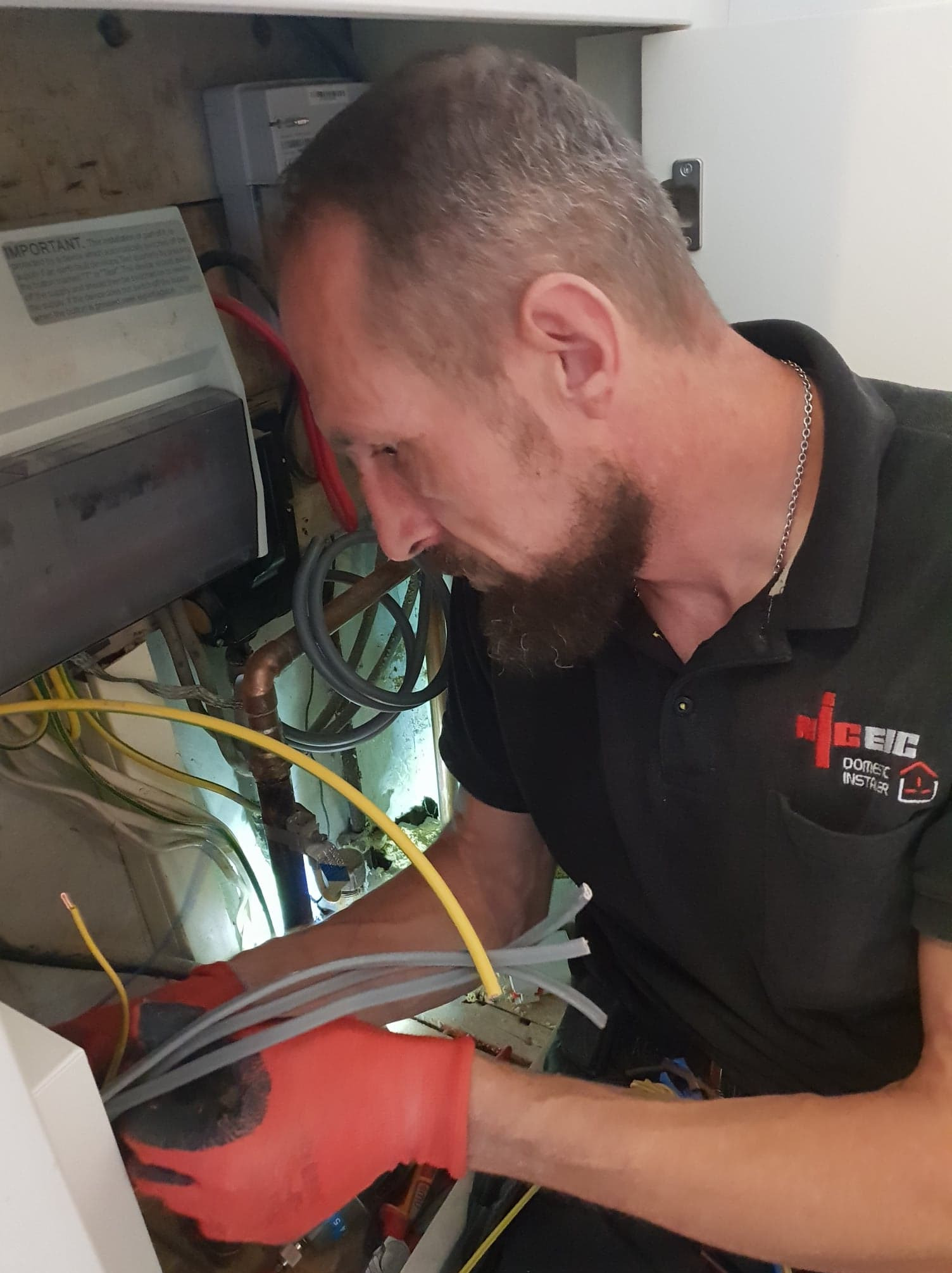 Elektryk w trakcie pracy nad instalacją elektryczną, w pomarańczowych rękawicach, z widocznymi przewodami i elementami skrzynki. Ujęcie z bliska, skupione na detalu.