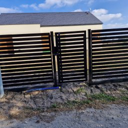 PRO FENCE KRZYSZTOF KOŁTYŚ - Nowoczesna, pozioma brama wjazdowa z czarnych, metalowych profili, widoczna z zewnątrz posesji, z fragmentem domu w tle i niebieskim karbowanym przewodem na ziemi.