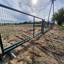 PRO FENCE KRZYSZTOF KOŁTYŚ - Nowo zamontowana, przesuwna brama z zielonej siatki panelowej, widoczna z bliska na tle piaszczystego terenu z wyraźnymi śladami opon, betonowy fundament i drewniane podpory pod bramą, słoneczny...