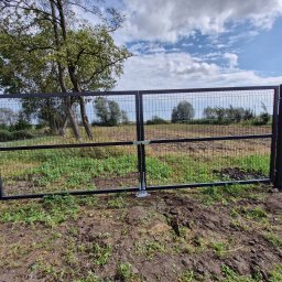 PRO FENCE KRZYSZTOF KOŁTYŚ - Nowoczesna, czarna, dwuskrzydłowa furtka z siatki z mechanizmem zamykającym, wkomponowana w ogrodzenie z siatki na tle zieleni i błękitnego nieba z chmurami.