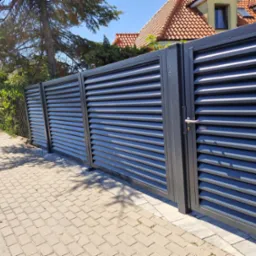 Nowoczesna, pozioma, metalowa brama wjazdowa i ogrodzenie w kolorze grafitowym, z widoczną klamką, zamontowane na posesji z kostką brukową, w tle dom z czerwoną dachówką i drzewa.