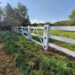 Nowo zainstalowane, pomalowane na biało ogrodzenie panelowe z poprzecznymi belkami, czarnymi zawiasami i widocznymi śladami montażu w postaci świeżej ziemi.