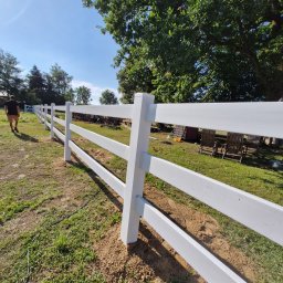 PRO FENCE KRZYSZTOF KOŁTYŚ - Nowe, białe, drewniane ogrodzenie rancho z trzema poprzeczkami, widok pod kątem, świeżo usypana ziemia wokół słupków, osoba w tle, drzewa i niebieskie niebo.