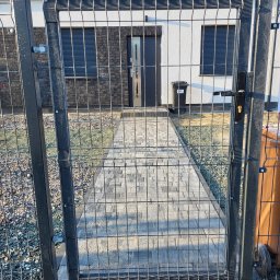 PRO FENCE KRZYSZTOF KOŁTYŚ - Widok przez otwartą, metalową furtkę z siatki na posesję z brukowanym chodnikiem prowadzącym do drzwi domu. W tle ściana domu z dwoma oknami z roletami zewnętrznymi.