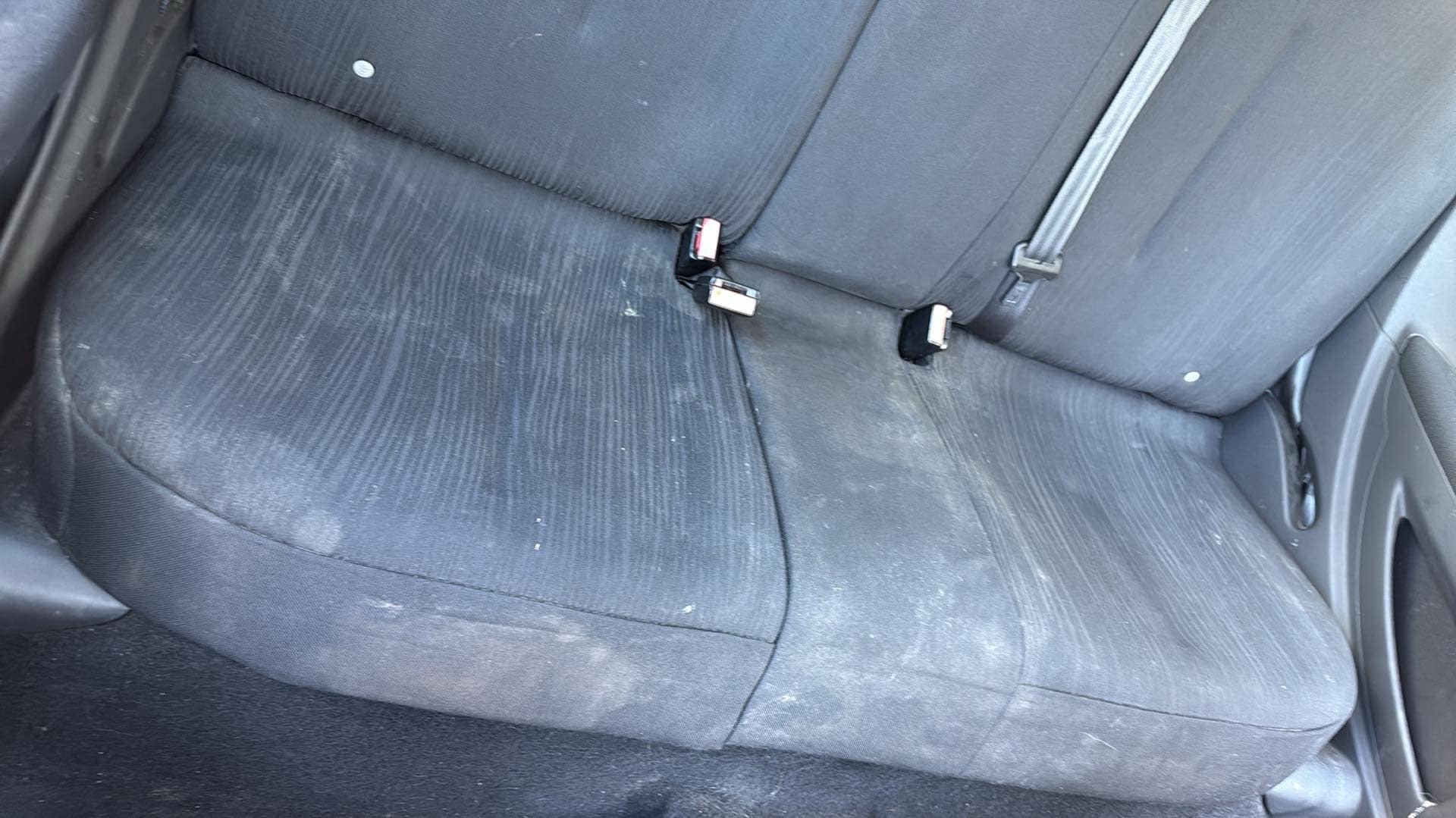 Szare, mocno zabrudzone siedzenie samochodowe z pasami bezpieczeństwa i widocznymi plamami na tapicerce.