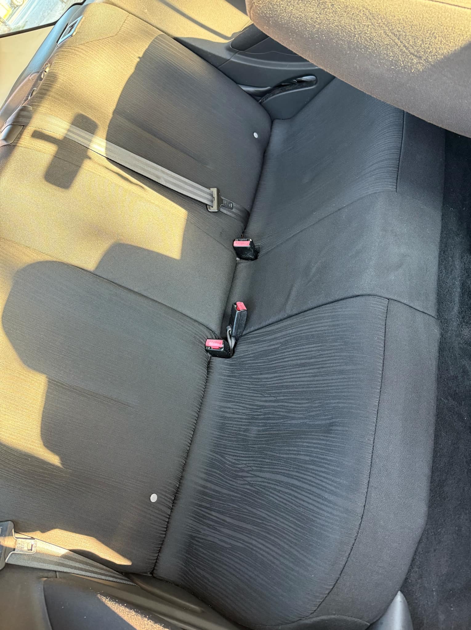Czarne, teksturowane tylne siedzenie samochodowe z widocznymi pasami bezpieczeństwa i metalowym elementem mocującym, oświetlone promieniami słonecznymi.