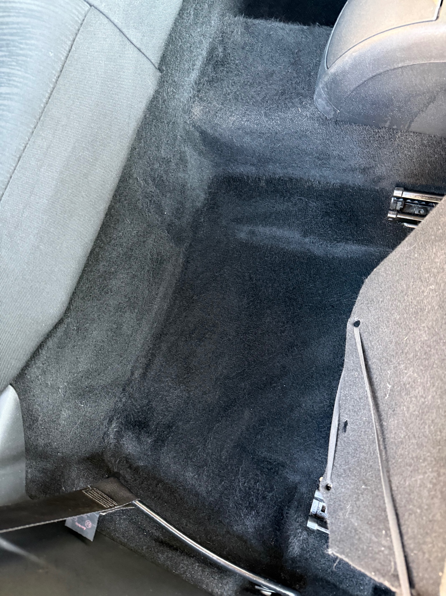 Ciemna, welurowa podłoga samochodu, widoczny fragment siedzenia i mocowania, z wyraźną różnicą w czystości między obszarem wyczyszczonym a zabrudzonym.