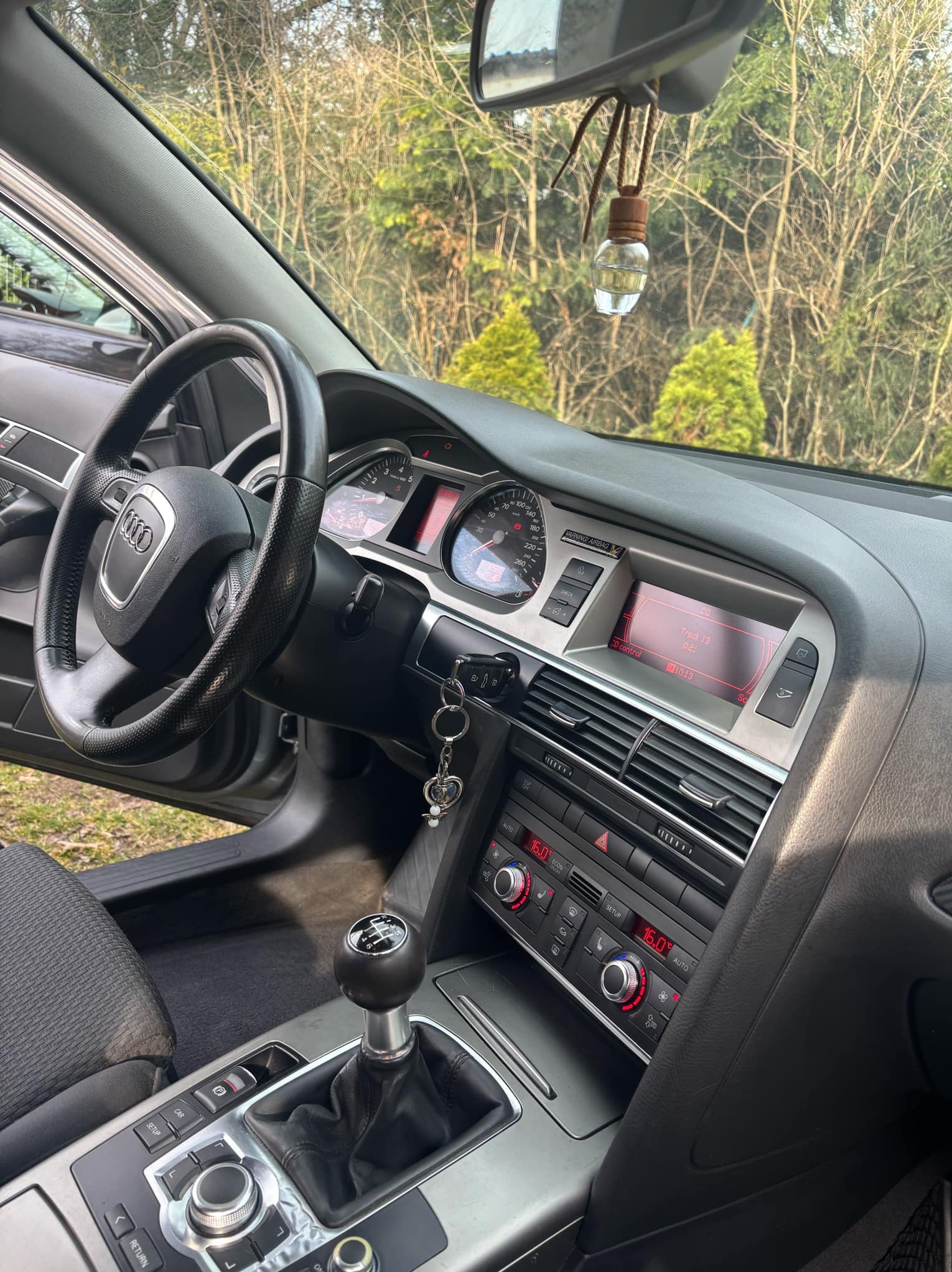 Wnętrze samochodu marki Audi z widoczną deską rozdzielczą, kierownicą, dźwignią zmiany biegów i wiszącym odświeżaczem powietrza w kształcie buteleczki.