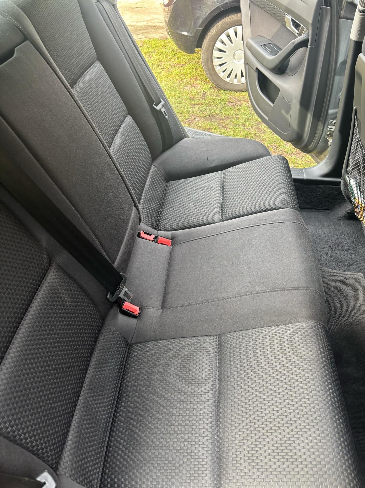 Wnętrze samochodu: tylna kanapa z ciemną tapicerką o wyraźnej fakturze, widoczne pasy bezpieczeństwa z czerwonymi zatrzaskami, fragment otwartych drzwi i koło samochodu na zewnątrz.