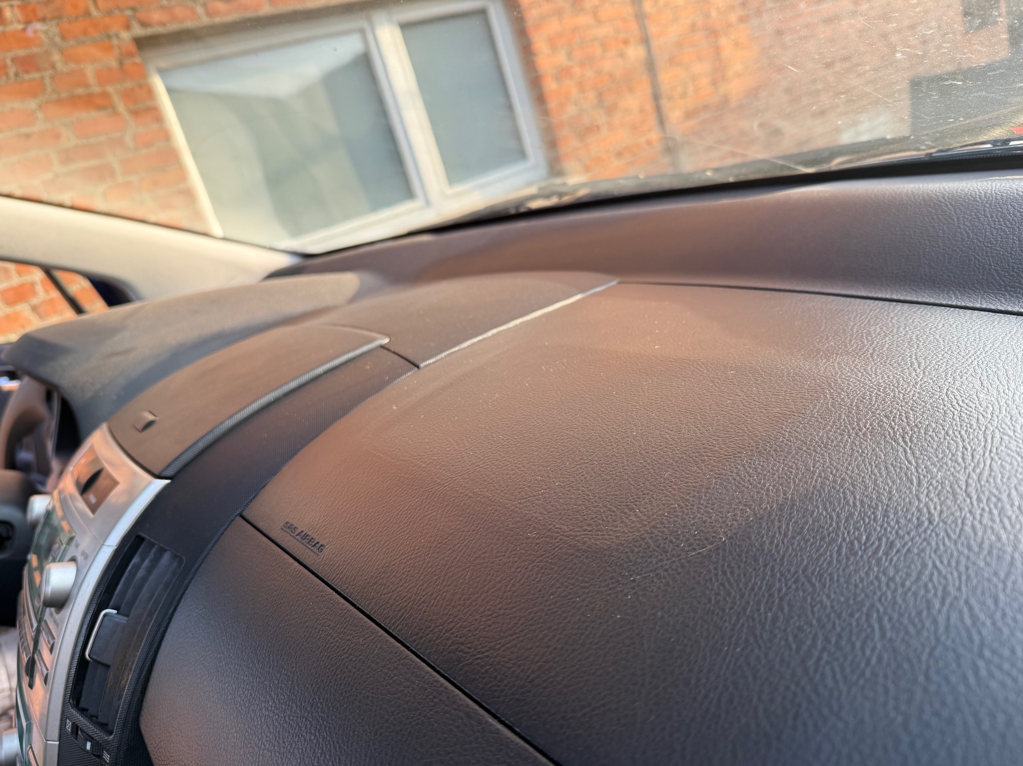 Szczegółowe ujęcie deski rozdzielczej samochodu w kolorze ciemnoszarym, z widocznymi detalami tekstury i napisem 'SRS AIRBAG', z fragmentem konsoli środkowej po lewej stronie i oknem z ceglanym...