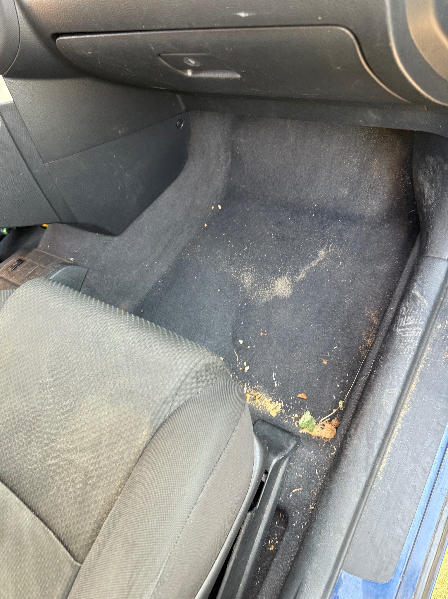 Wnętrze samochodu z widocznym brudnym dywanikiem i fotelem kierowcy, zanieczyszczone piaskiem, okruchami i liśćmi.