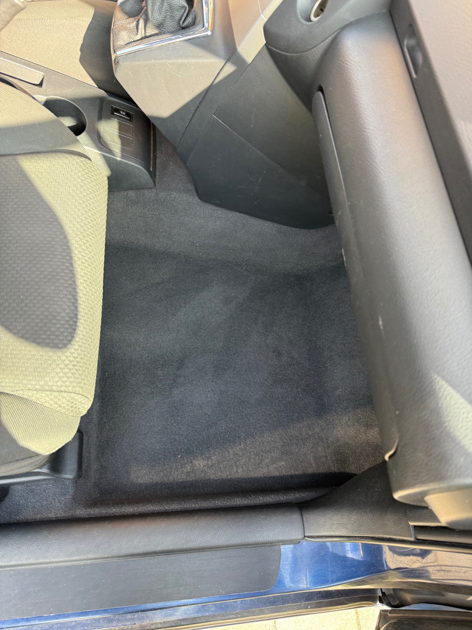 Wnętrze samochodu z wyczyszczoną tapicerką podłogową, widoczny fragment fotela, konsola środkowa i próg drzwi.