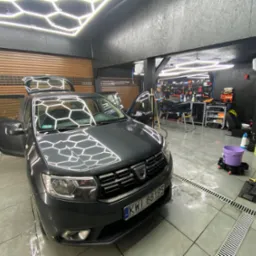 Szary samochód Dacia z otwartymi drzwiami i bagażnikiem w myjni z podświetleniem sufitowym w kształcie plastra miodu, widoczny pracownik w tle.