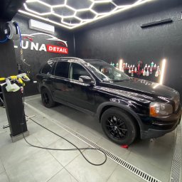 Luna Detail - Czarny SUV marki Volvo na podnośnikach w nowoczesnej myjni detailingowej z oświetleniem LED w kształcie plastra miodu i logo 'Luna Detail' na ścianie.