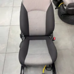 Wyjęte z samochodu, czyste, szaro-czarne siedzenie samochodowe z pasami bezpieczeństwa, ustawione na szarej podłodze z płytek. W tle widoczne opony i dywaniki samochodowe.