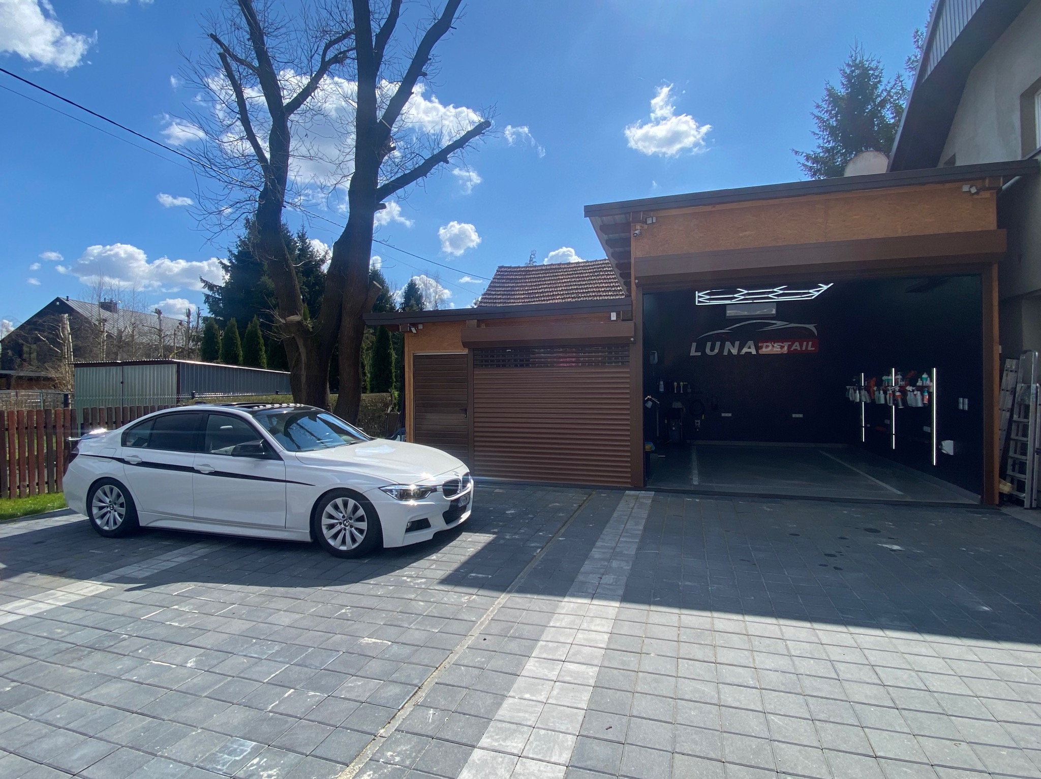 Białe BMW z czarnymi pasami na tle garażu Luna Detail w Kłaju, w tle drzewa i błękitne niebo z chmurami.