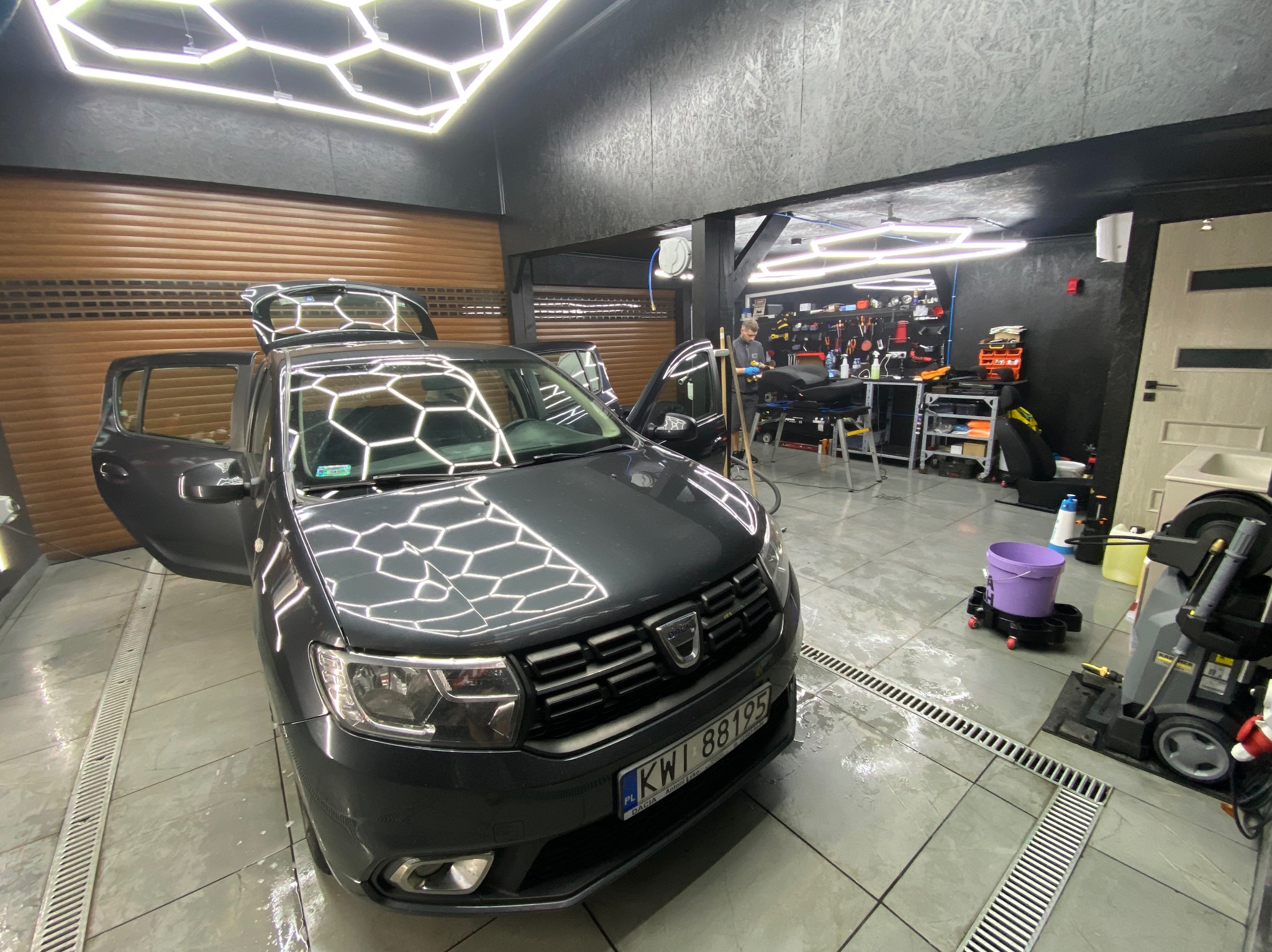 Szary samochód Dacia z otwartymi drzwiami i bagażnikiem w myjni z podświetleniem sufitowym w kształcie plastra miodu, widoczny pracownik w tle.