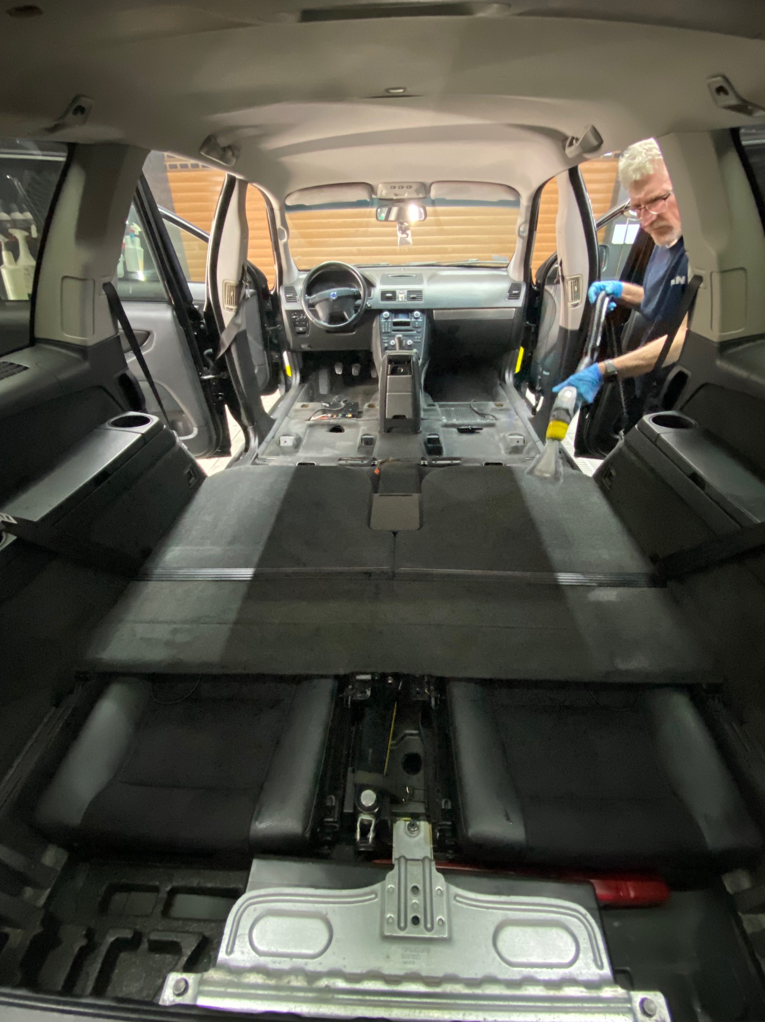 Wnętrze samochodu Volvo XC90 w trakcie czyszczenia tapicerki podłogowej za pomocą odkurzacza piorącego przez mężczyznę w okularach i niebieskich rękawicach, z widoczną deską rozdzielczą i otwartymi...