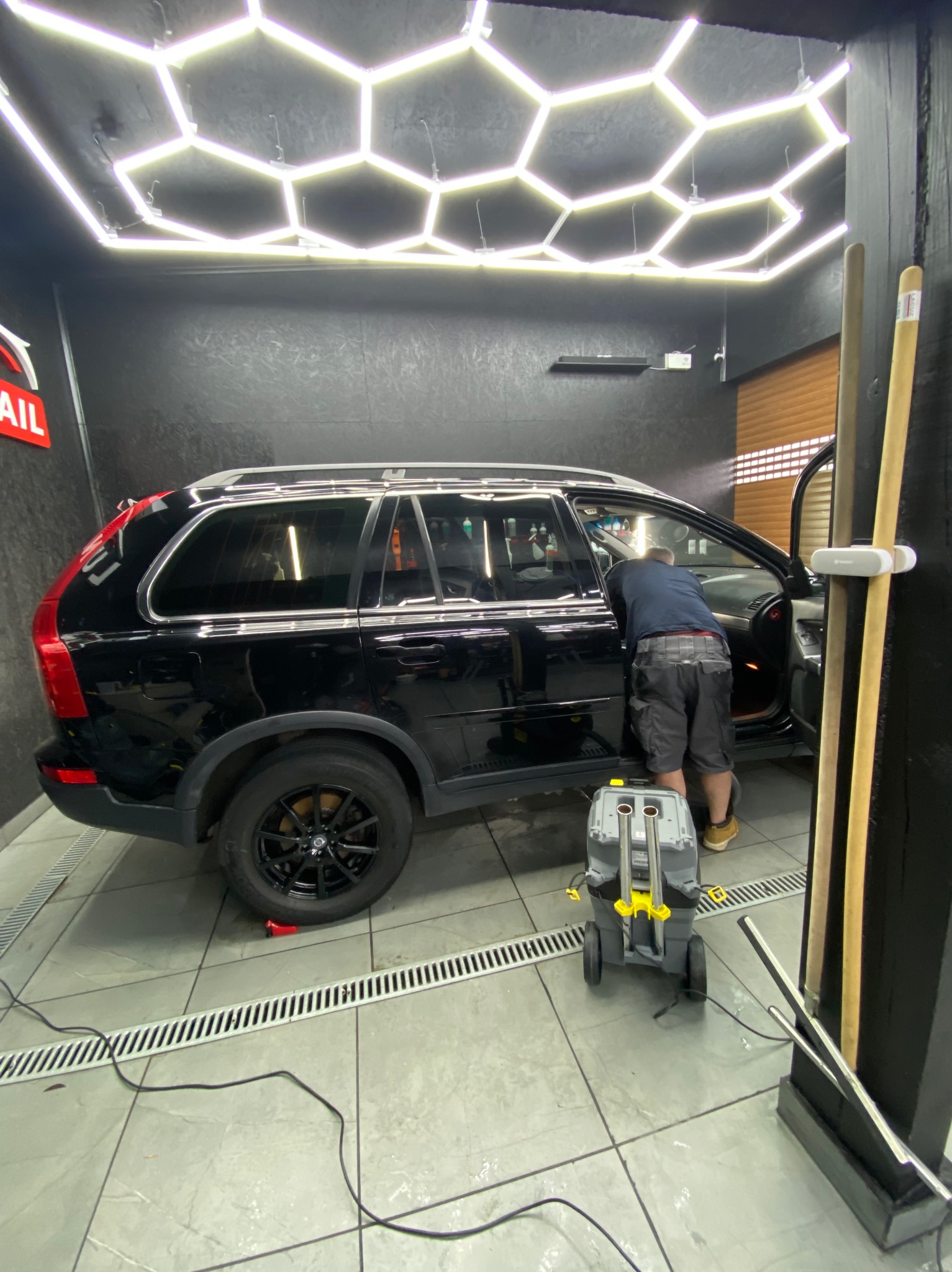 Czarny SUV w myjni z oświetleniem LED w kształcie plastra miodu; mężczyzna w niebieskiej koszulce czyści wnętrze, obok stoi maszyna czyszcząca.