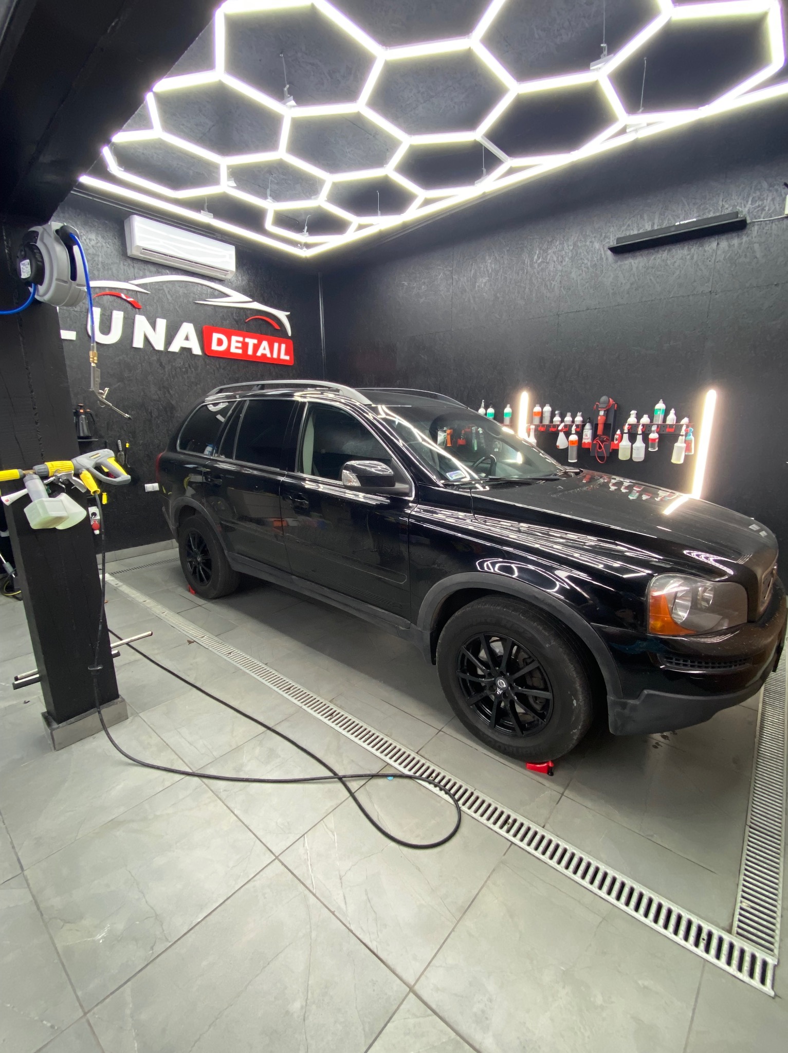 Czarny SUV marki Volvo na podnośnikach w nowoczesnej myjni detailingowej z oświetleniem LED w kształcie plastra miodu i logo 'Luna Detail' na ścianie.