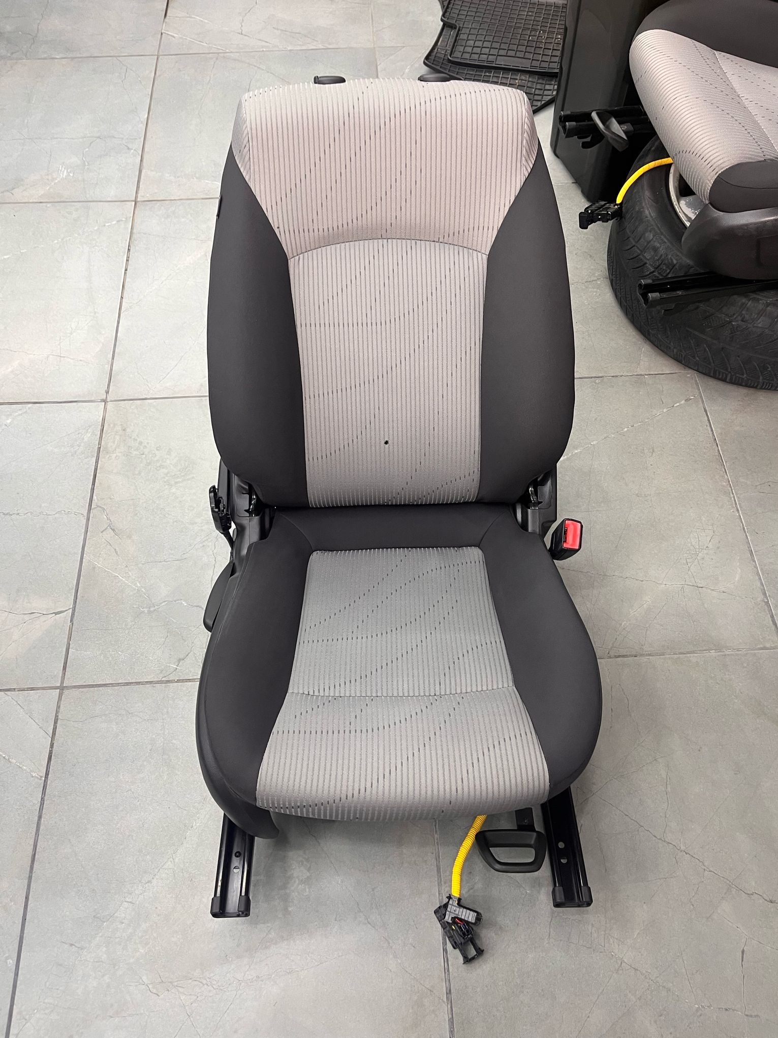 Wyjęte z samochodu, czyste, szaro-czarne siedzenie samochodowe z pasami bezpieczeństwa, ustawione na szarej podłodze z płytek. W tle widoczne opony i dywaniki samochodowe.