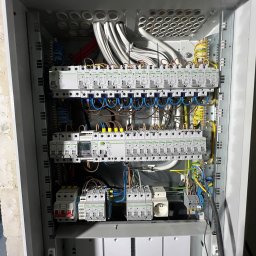 Maksymilian Ficht - Otwarta szara skrzynka rozdzielcza z widocznymi bezpiecznikami, przewodami i innymi elementami instalacji elektrycznej, zamontowana na ścianie z surowego betonu.