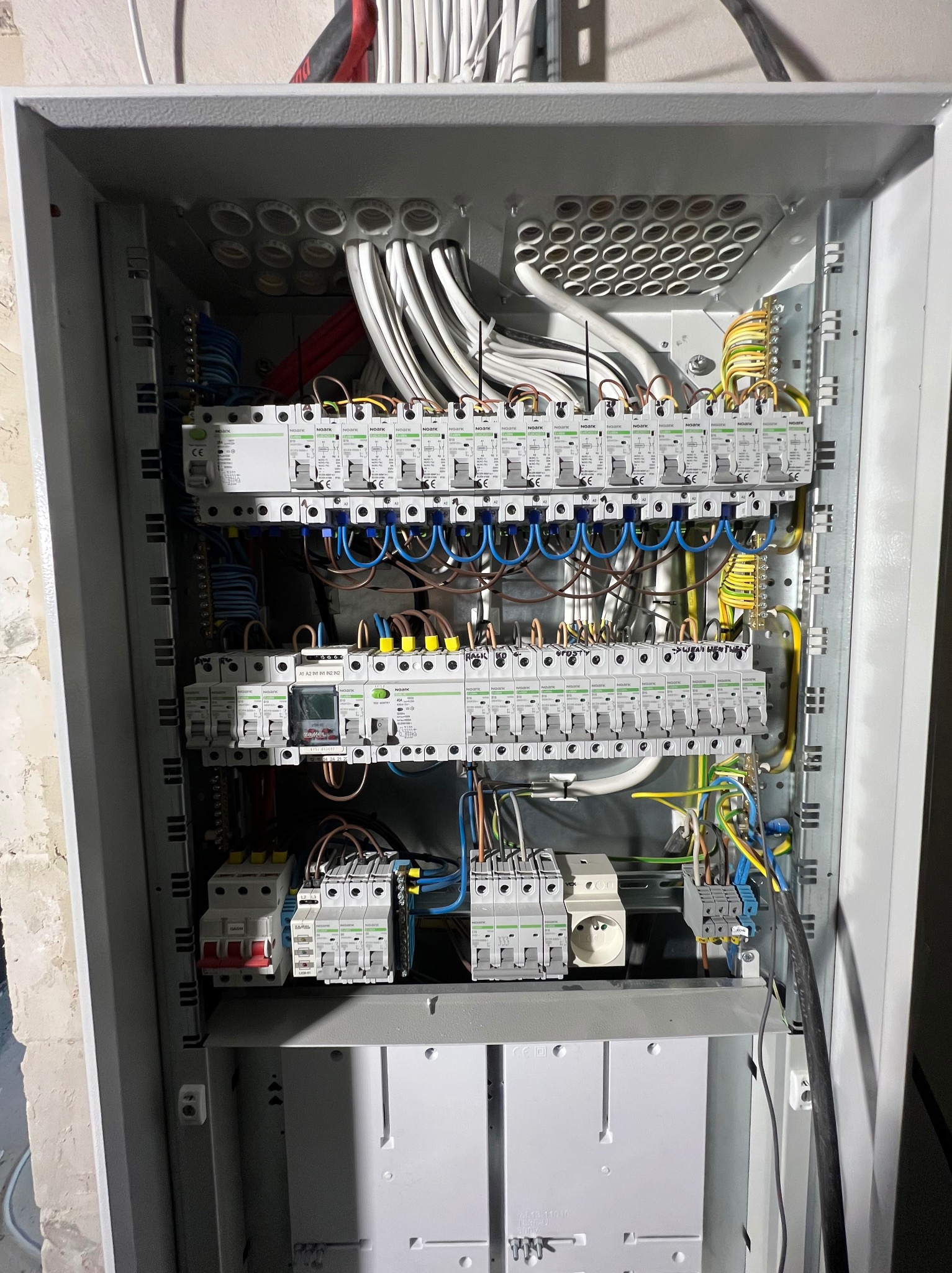 Otwarta szara skrzynka rozdzielcza z widocznymi bezpiecznikami, przewodami i innymi elementami instalacji elektrycznej, zamontowana na ścianie z surowego betonu.