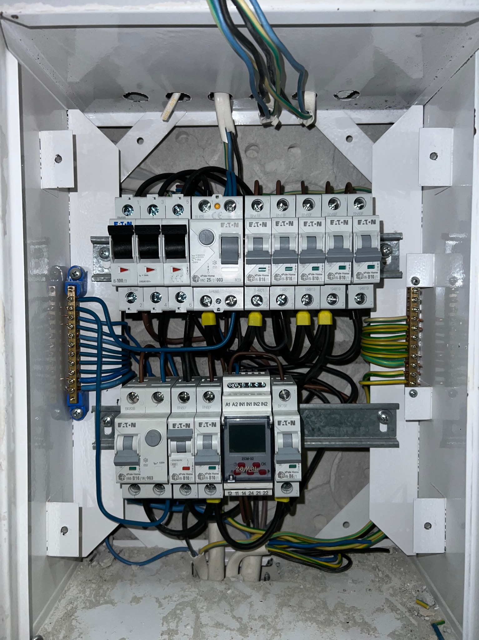 Otwarta skrzynka rozdzielcza z zamontowanymi bezpiecznikami i przewodami, widoczne elementy marki Eaton, instalacja w trakcie prac, widoczne ślady kurzu i zabrudzeń.