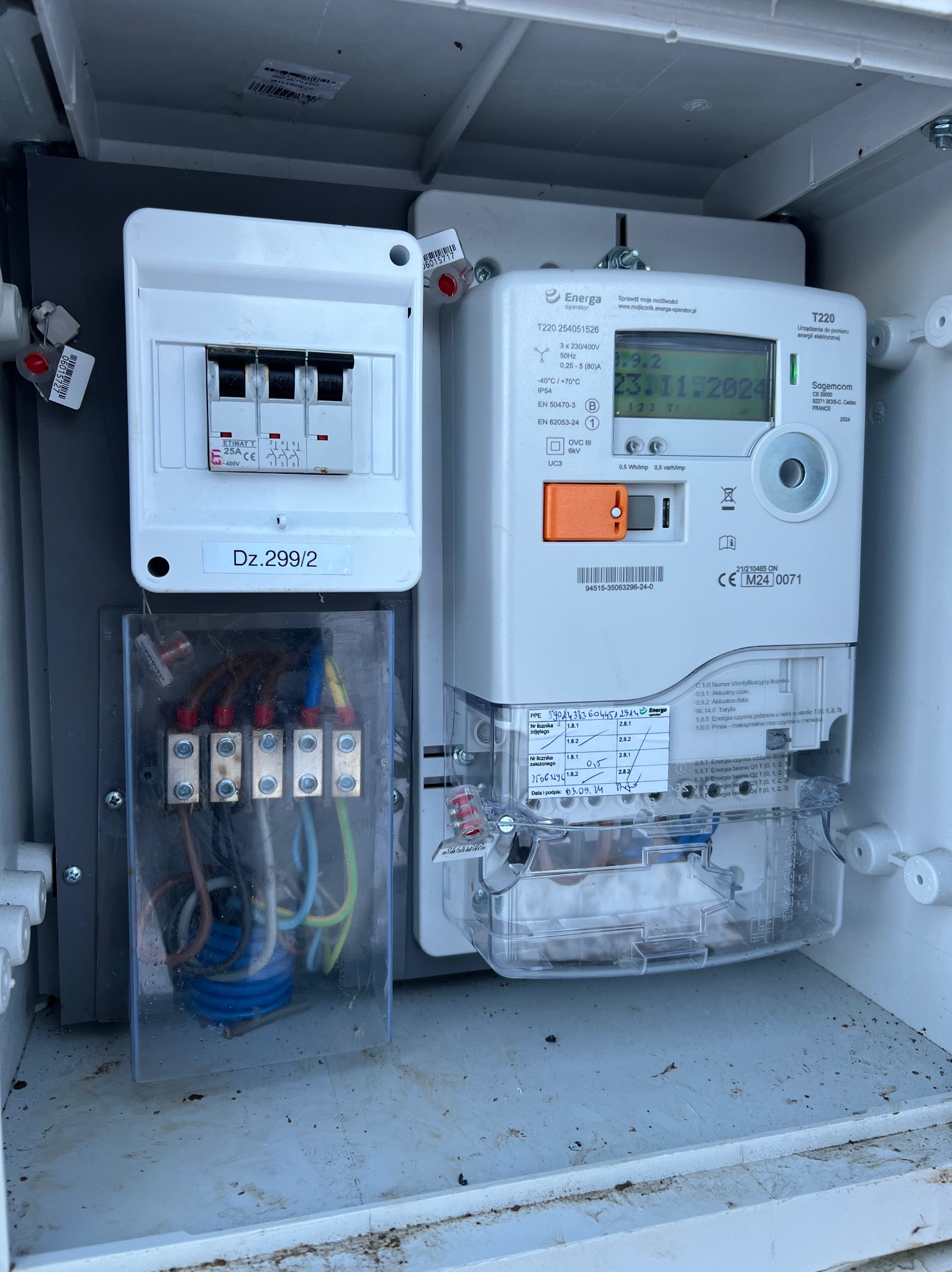 Otwarta skrzynka elektryczna z licznikiem Energii, bezpiecznikami i złączami kablowymi, widoczne przewody w kolorach brązowym, niebieskim i żółto-zielonym.