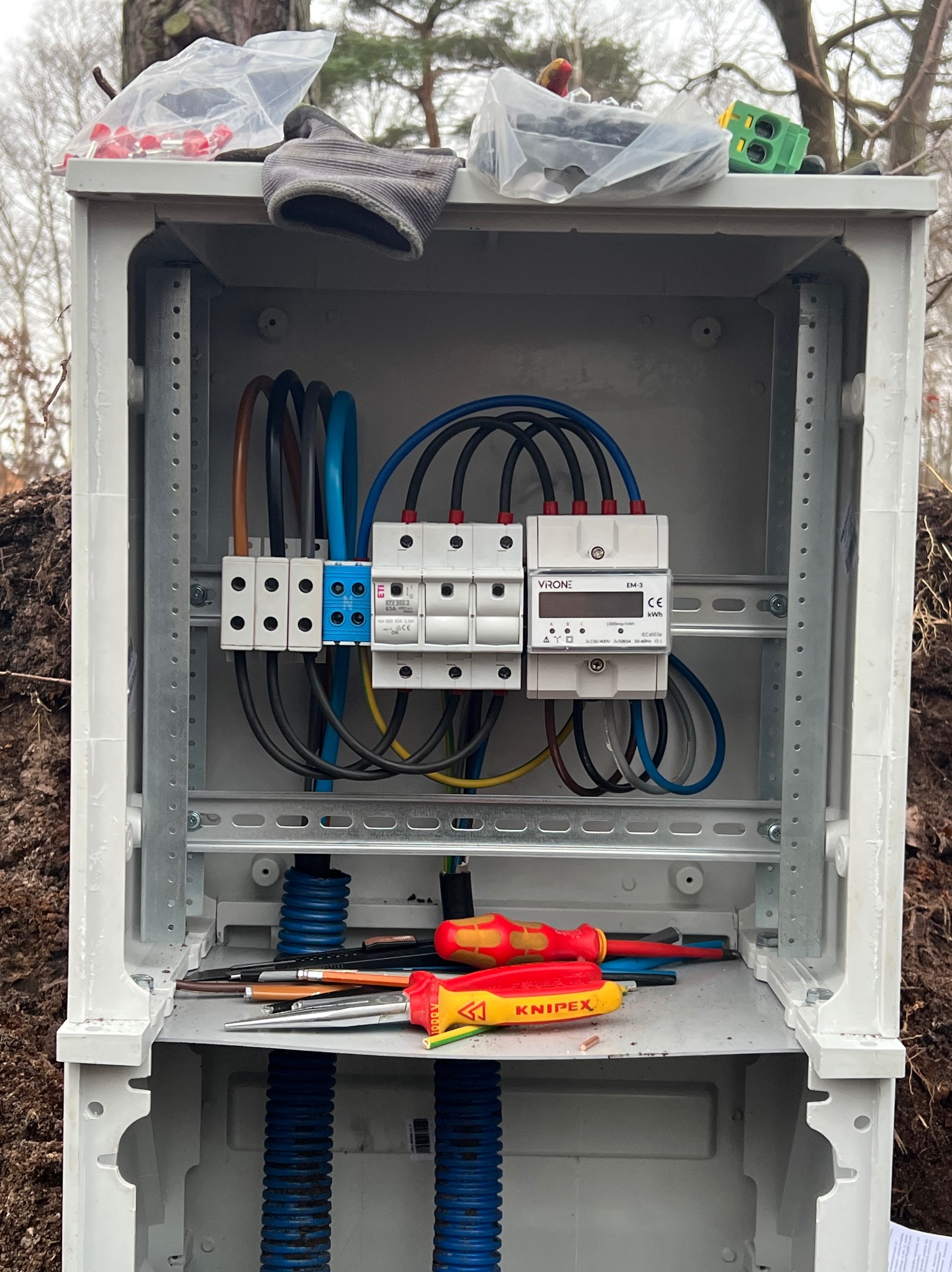 Otwarta skrzynka elektryczna z zainstalowanymi bezpiecznikami, licznikiem energii i okablowaniem w Bolszewie. Na dnie skrzynki leżą narzędzia, w tym śrubokręty i obcęgi marki Knipex.