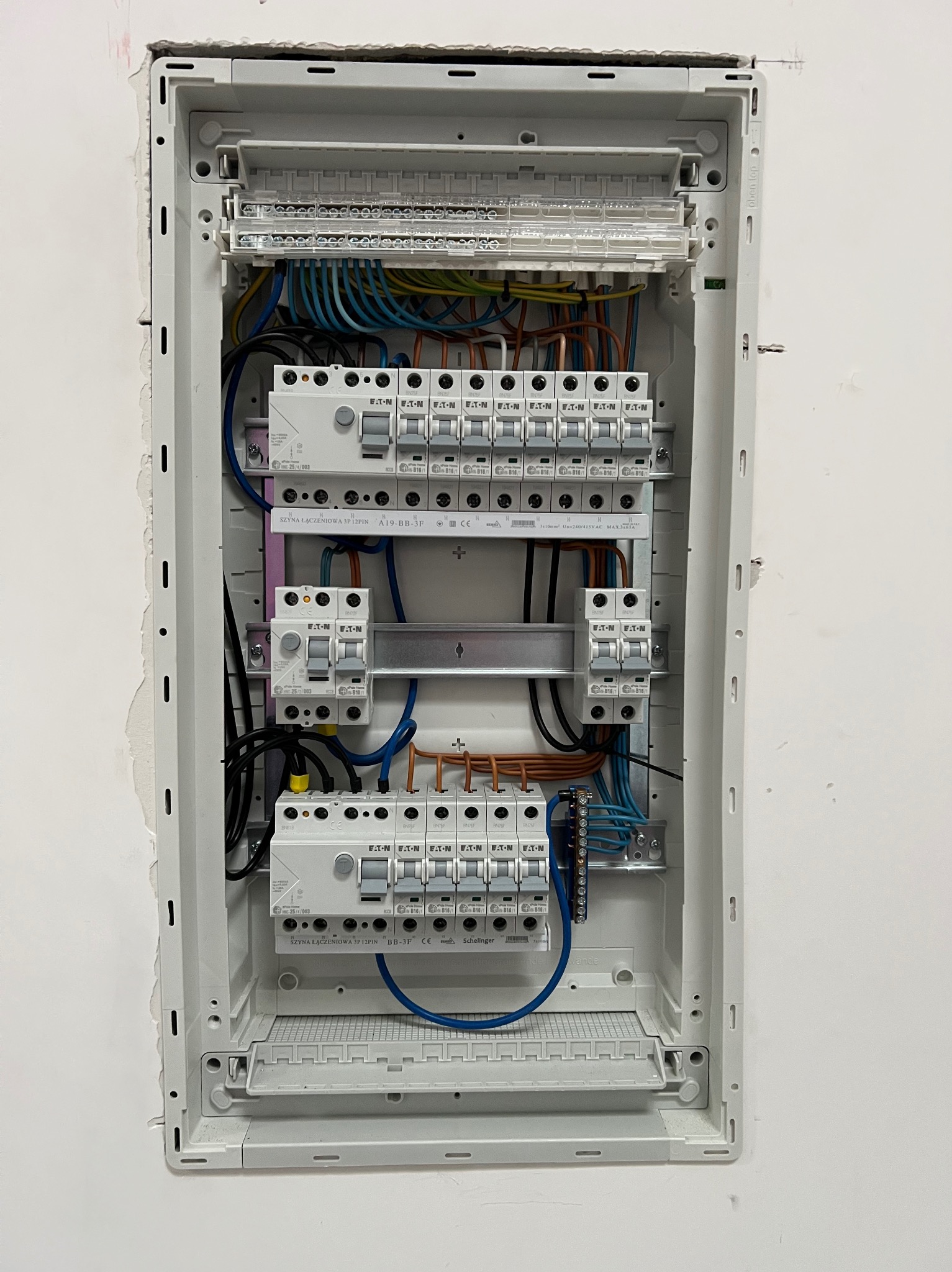 Rozdzielnica elektryczna wnękowa z zainstalowanymi bezpiecznikami, przewodami i szynami łączeniowymi, widoczne marki Eaton i Schelinger, montaż w ścianie z widocznymi śladami po instalacji.