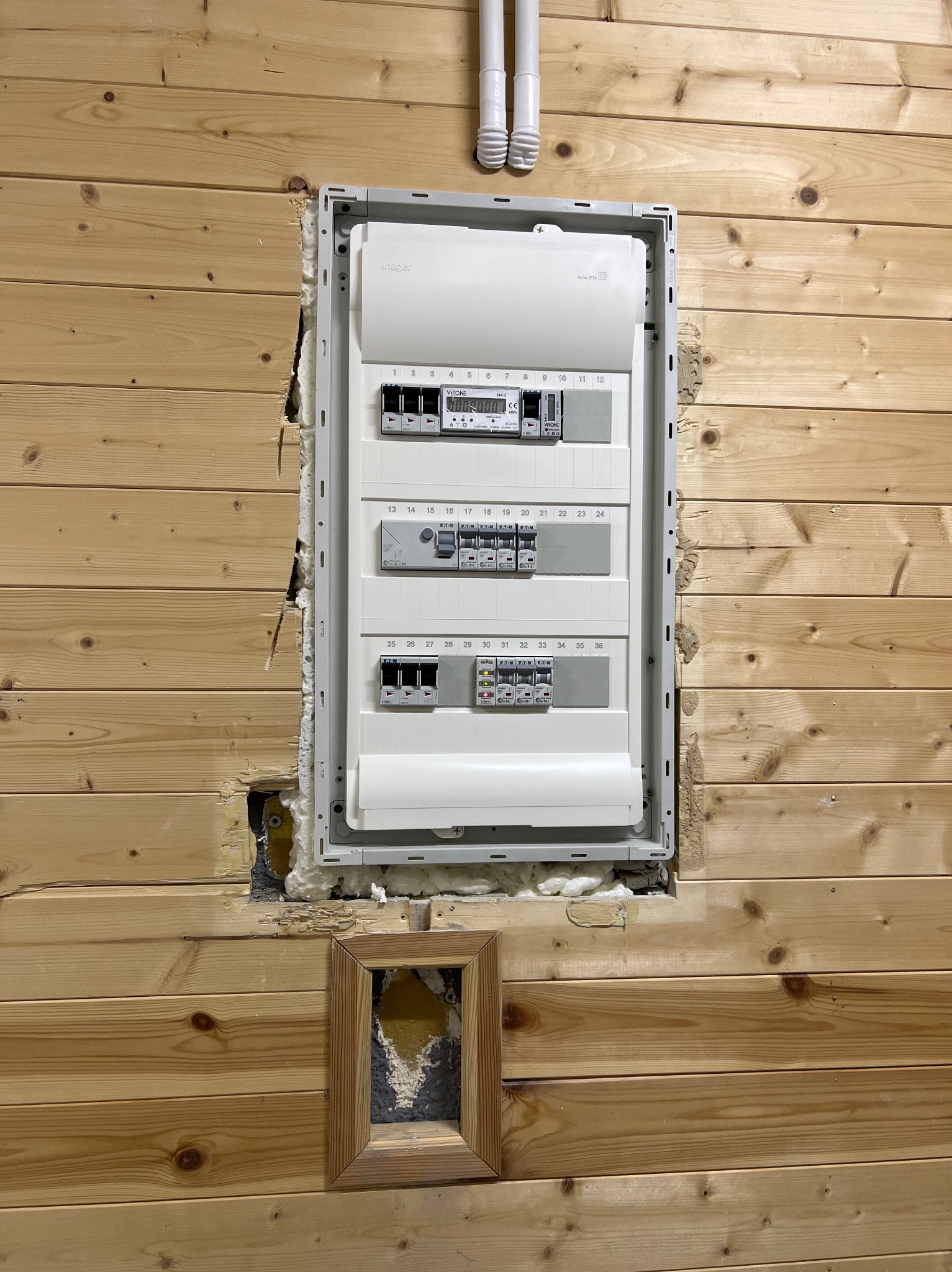 Rozdzielnica elektryczna zamontowana w drewnianej ścianie, widoczne niedoskonałości wykończenia wokół obudowy, szczeliny wypełnione pianką montażową, w tle rury instalacyjne i ramka po zdjęciu.