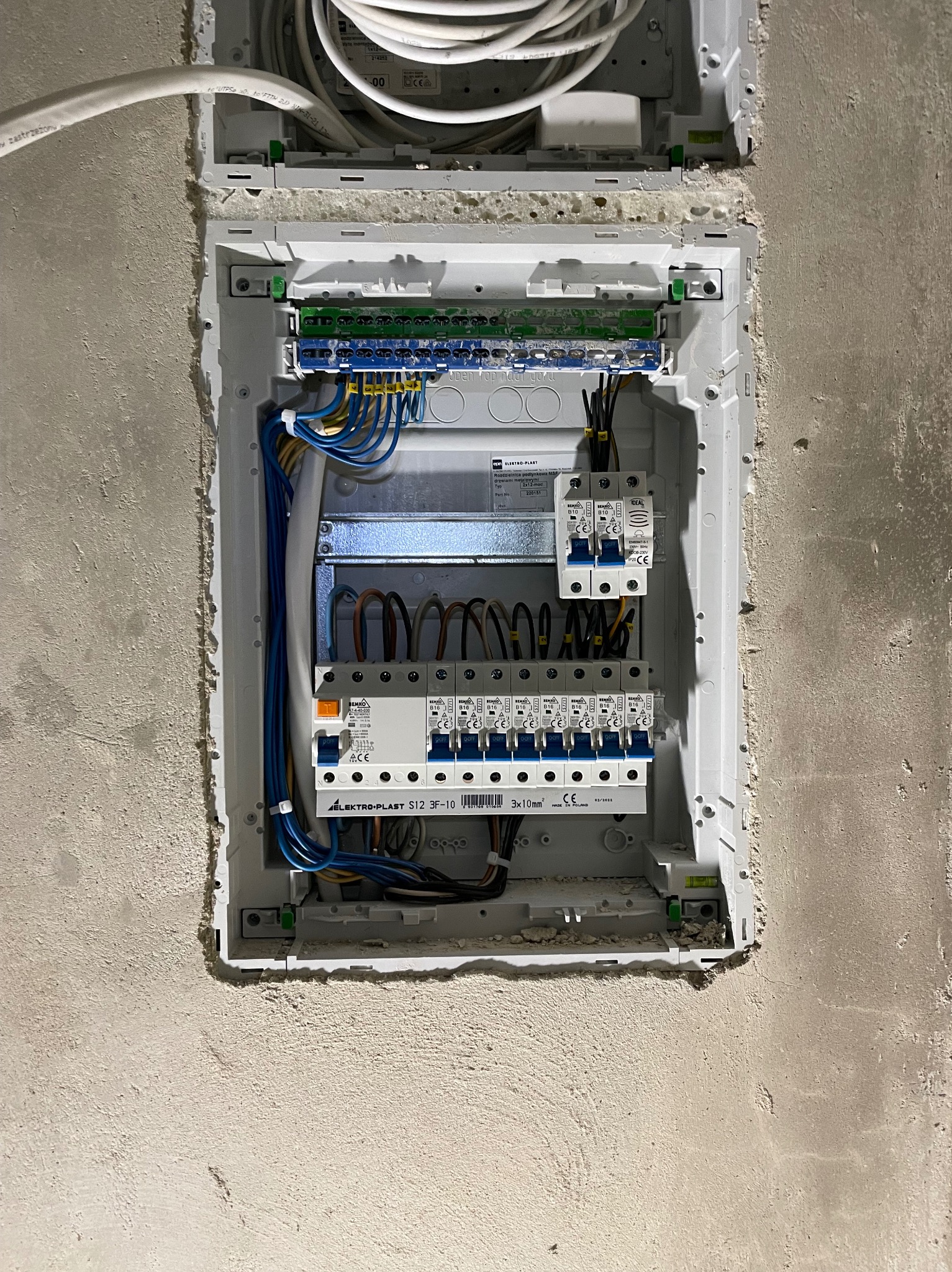 Otwarta skrzynka rozdzielcza z zamontowanymi bezpiecznikami i podłączonymi przewodami w surowej ścianie, widoczne oznaczenia producenta Elektro-Plast.