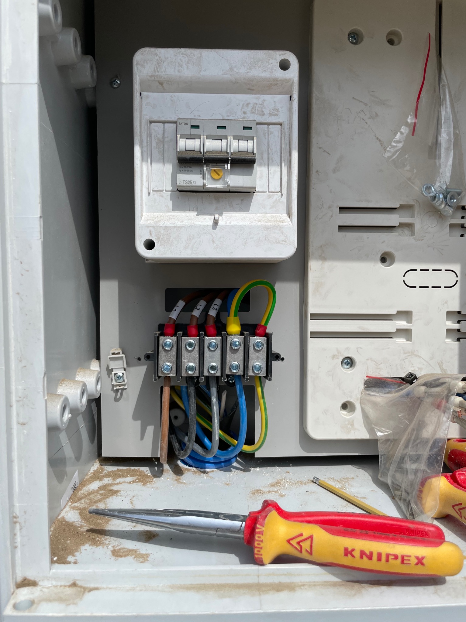 Wnętrze skrzynki elektrycznej z zamontowanymi bezpiecznikami EATON i podłączonymi przewodami oznaczonymi L1, L2, L3, widoczne złącza śrubowe, u dołu leży śrubokręt izolowany KNIPEX 1000V...