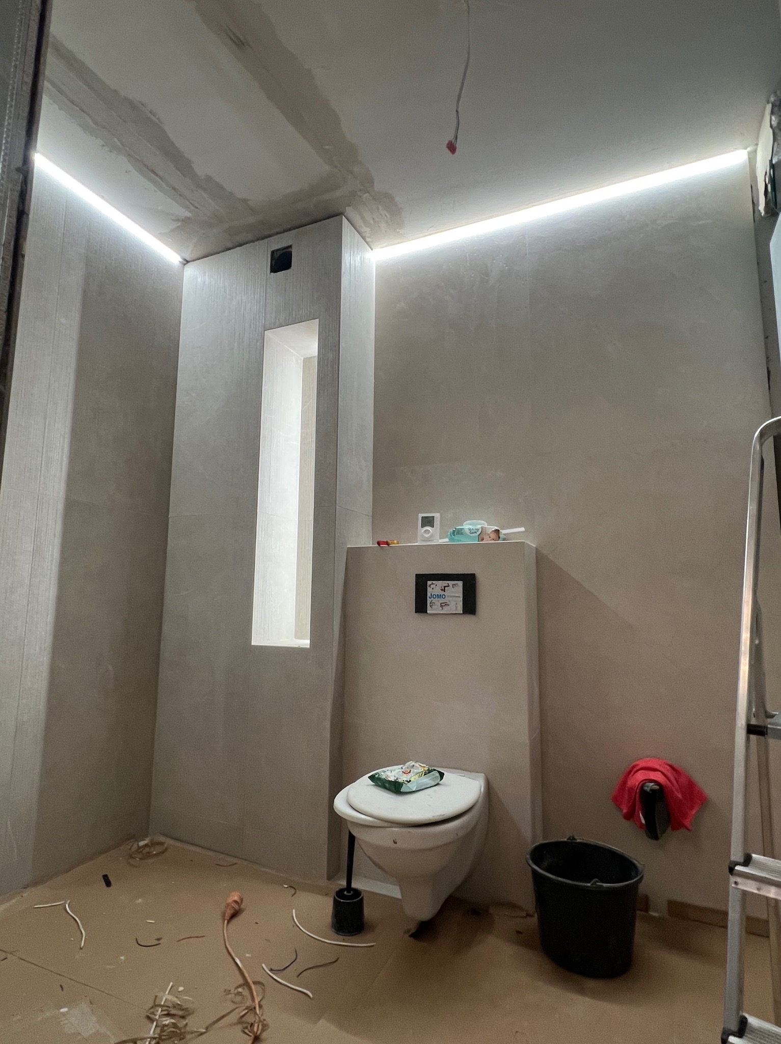 Wnętrze łazienki w trakcie remontu, widoczne podświetlenie LED w suficie, zabudowa toalety, niedokończone ściany i rozłożone kable na podłodze.