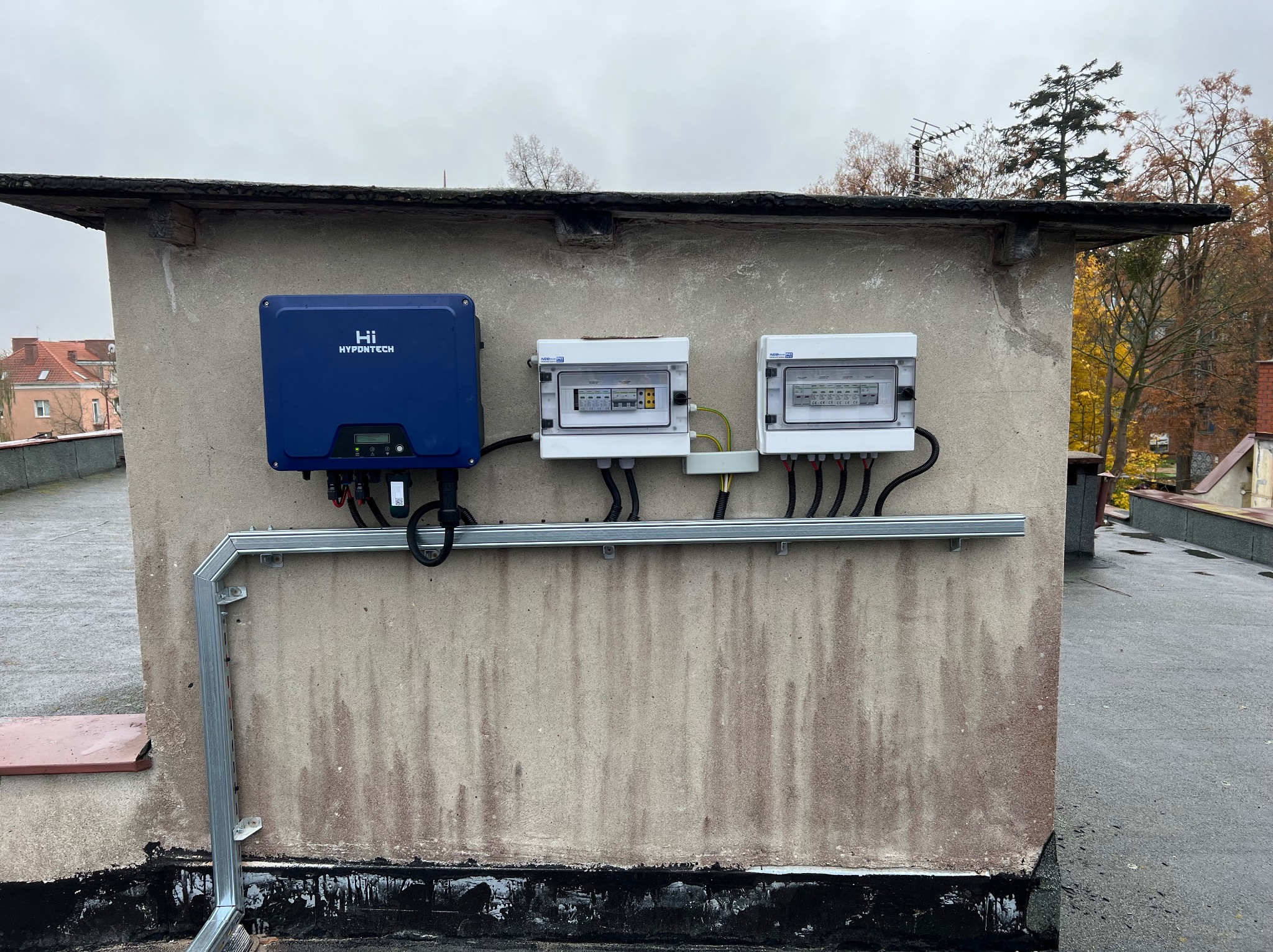 Instalacja fotowoltaiczna na dachu budynku: niebieski falownik Hypontech i dwie białe skrzynki rozdzielcze z przewodami, zamontowane na szarej, betonowej ścianie.