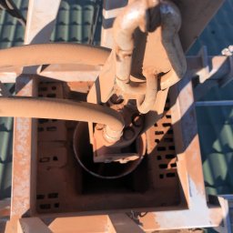 PROBI Kamil Kacprzak - Wnętrze komina z widocznymi elementami konstrukcyjnymi i przewodami, pokryte warstwą rdzawo-brązowego pyłu, na tle zielonego dachu.