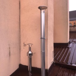 PROBI Kamil Kacprzak - Dwa chromowane przewody kominowe na dachu z blachy trapezowej w kolorze brązowym, w tle ściana budynku o nierównej strukturze.