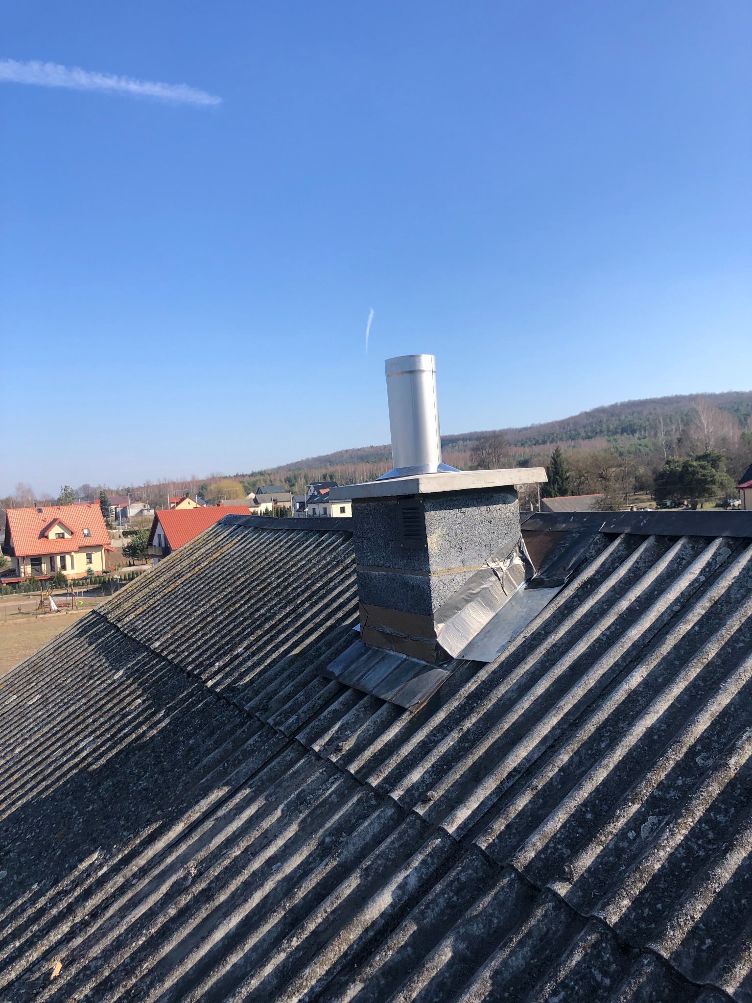 Nowy, srebrny wkład kominowy na szczycie murowanego komina na dachu z eternitu, widok na zabudowania i zalesione wzgórze w oddali.
