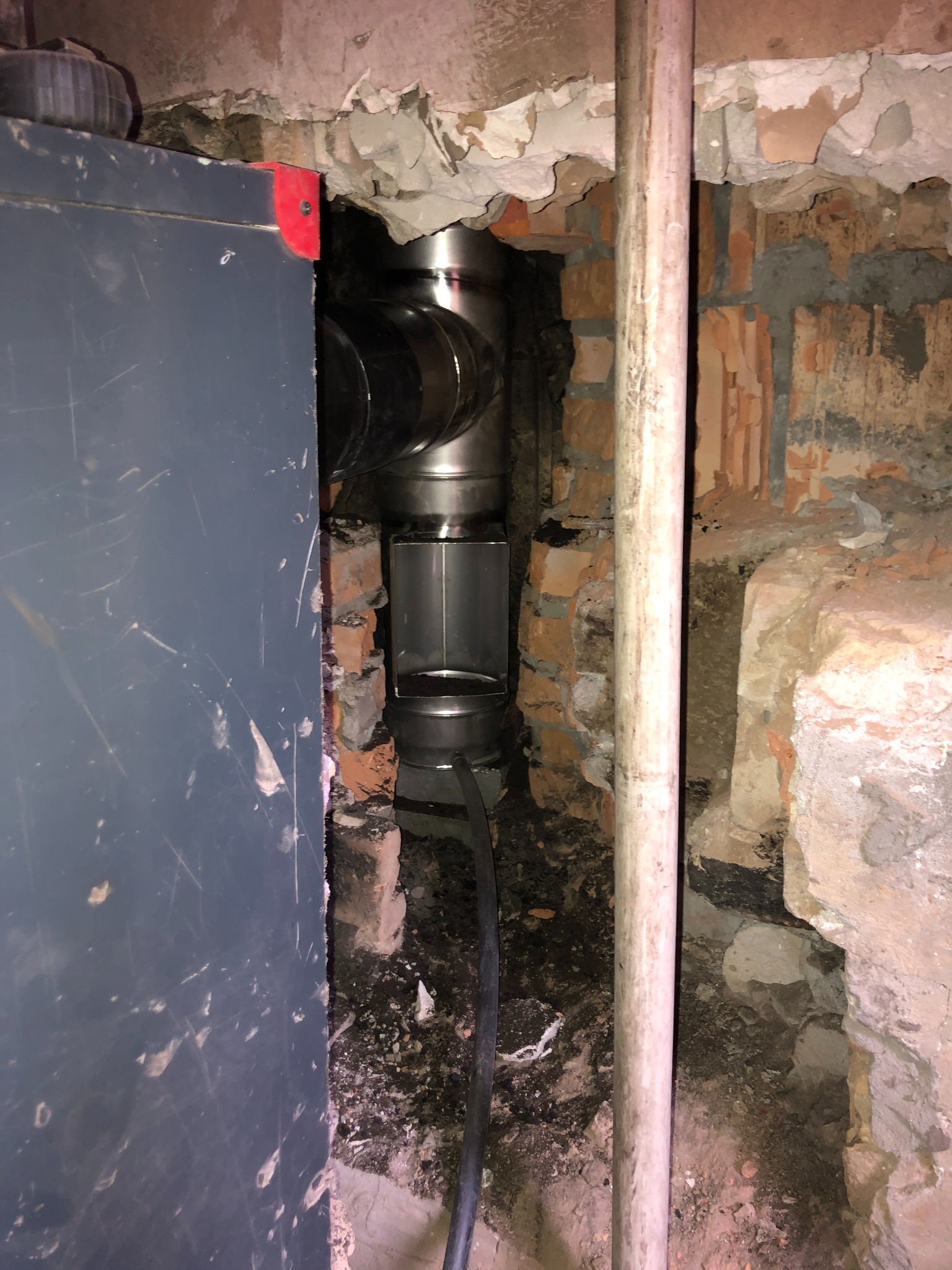 Fragment instalacji wentylacyjnej z metalowych rur w surowym, ceglanym wnętrzu, widoczny obok boku kotła CO.