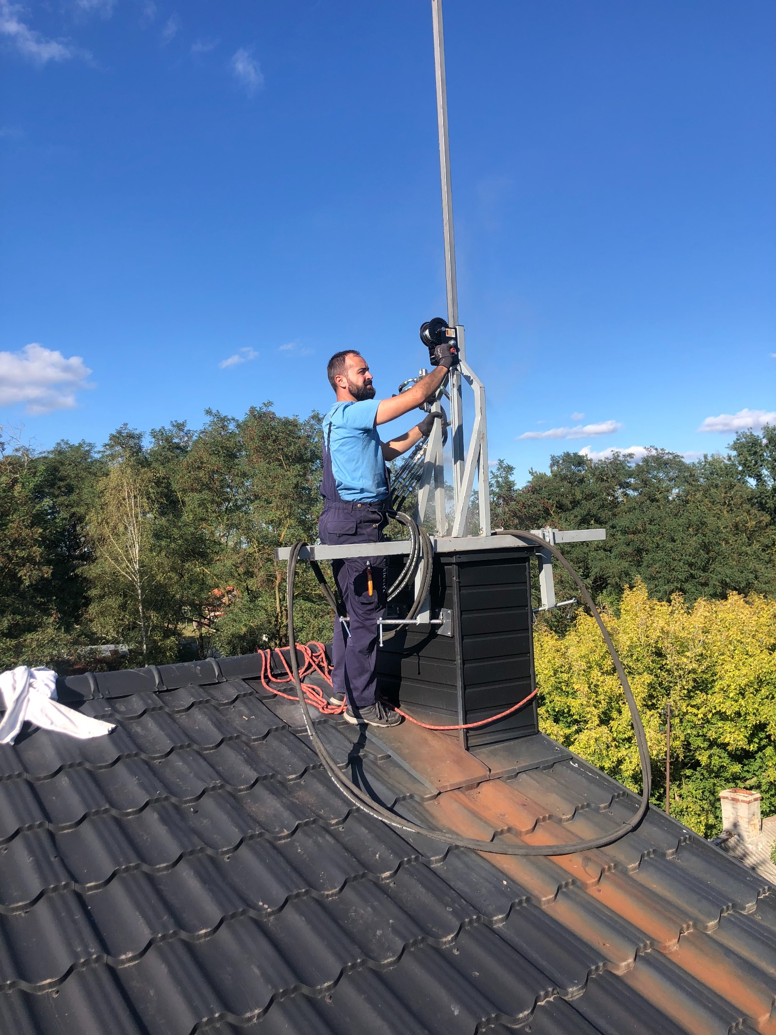 Kominiarz w niebieskim kombinezonie montuje odgromnik na czarnym kominie na tle błękitnego nieba.