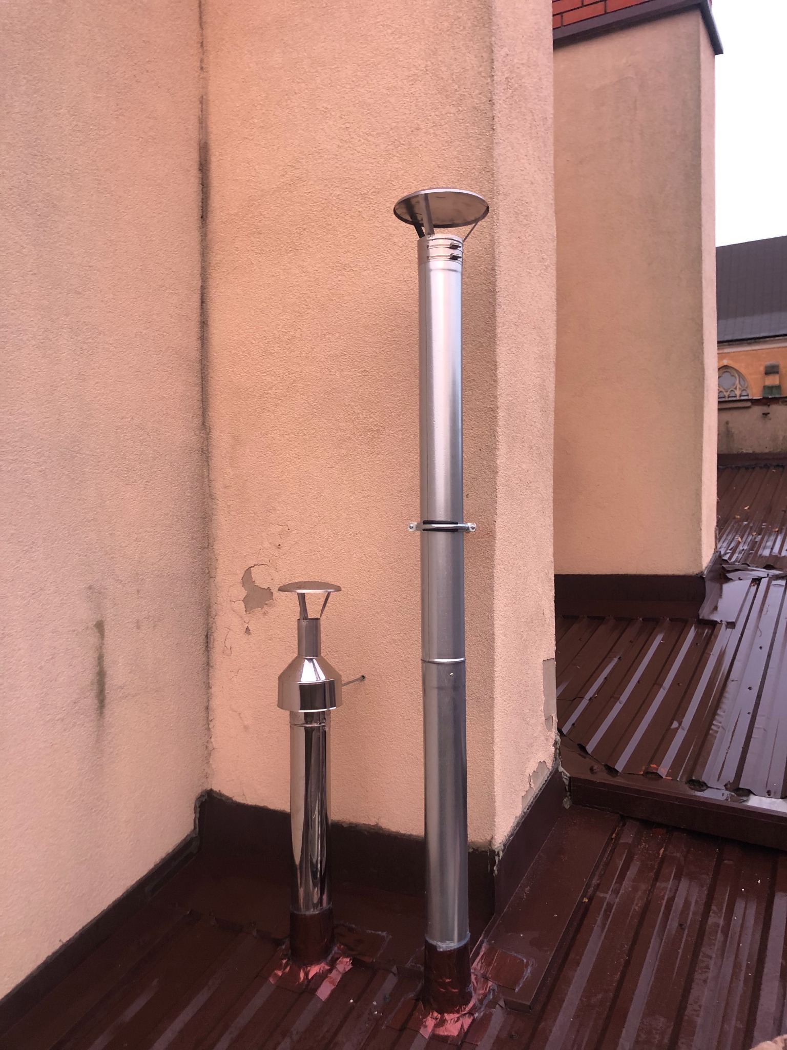 Dwa chromowane przewody kominowe na dachu z blachy trapezowej w kolorze brązowym, w tle ściana budynku o nierównej strukturze.