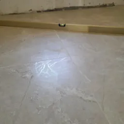 Położone jasne płytki podłogowe imitujące marmur, sprawdzane poziomicą na tle ściany w trakcie prac wykończeniowych.