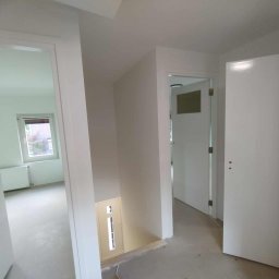 SZCZYRKBUD GRZEGORZ SOBAŃSKI, MAGDA KRUPIŃSKA SPÓŁKA CYWILNA - Białe, świeżo pomalowane wnętrze mieszkania z otwartymi drzwiami, widok na pokój z oknem. Podłoga w trakcie prac wykończeniowych.