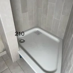 Narożny brodzik z odpływem w kabinie prysznicowej wyłożonej szarymi, pionowymi płytkami. Minimalistyczny design, jasne wnętrze łazienki.