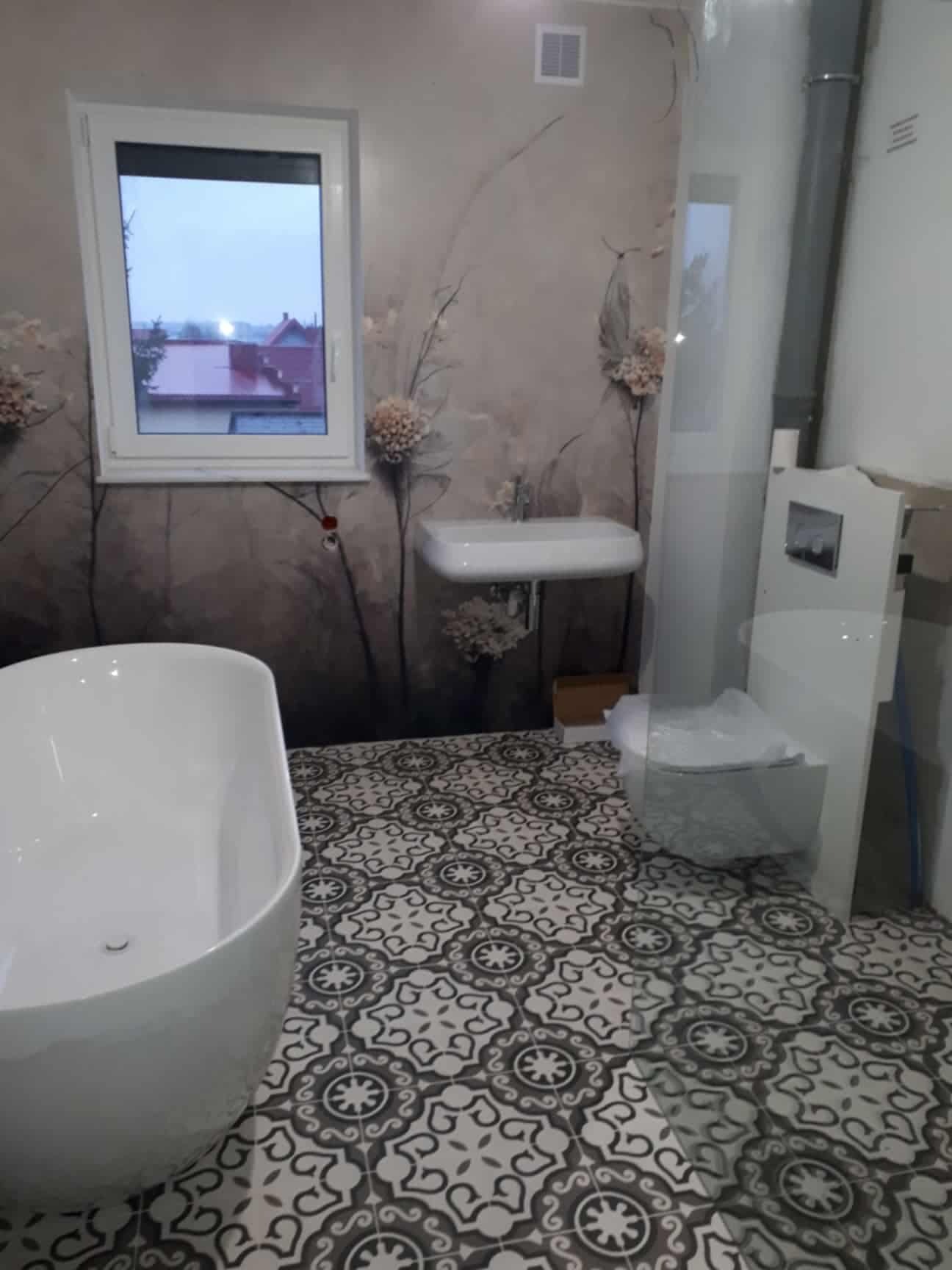 Nowoczesna łazienka z wolnostojącą wanną, umywalką i podwieszaną toaletą. Na ścianie tapeta w kwiaty, na podłodze wzorzyste płytki. Widok z okna na zewnątrz.