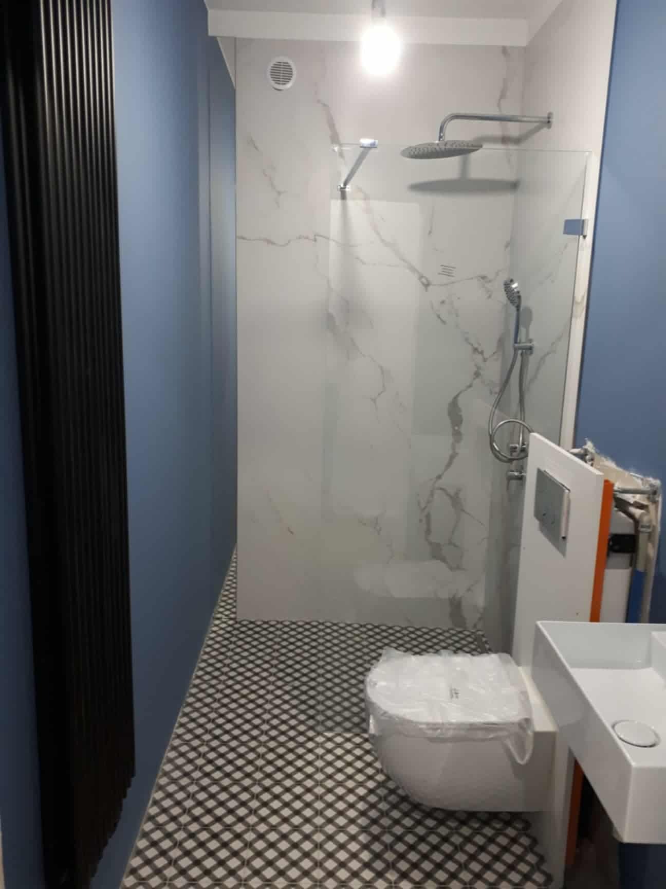 Nowoczesna łazienka po remoncie: kabina typu walk-in z deszczownicą, białe płytki imitujące marmur, czarno-biała podłoga w kratkę, niebieskie ściany i czarny grzejnik.