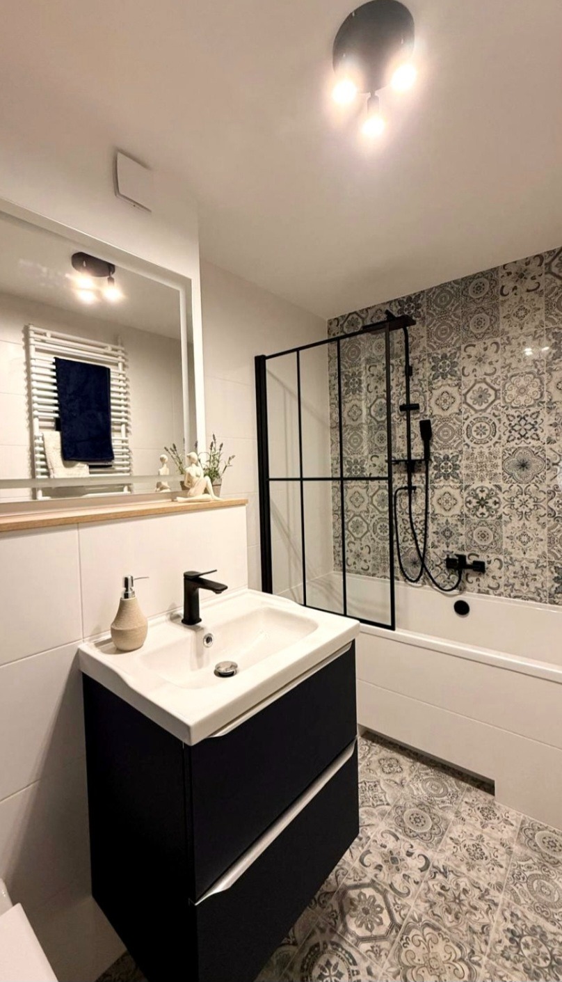 Nowoczesna łazienka z czarną armaturą, wzorzystymi płytkami i kabiną prysznicową typu walk-in. Minimalistyczna umywalka z szafką i lustro z oświetleniem.